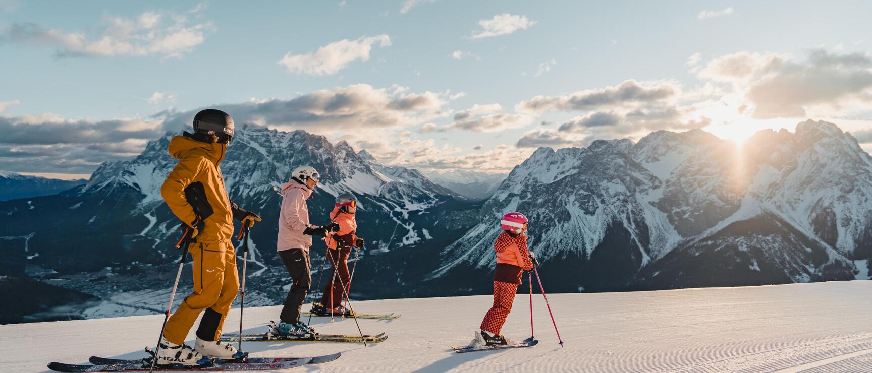 Four skiers on groomed slope with snow-covered mountain range and sun in background.