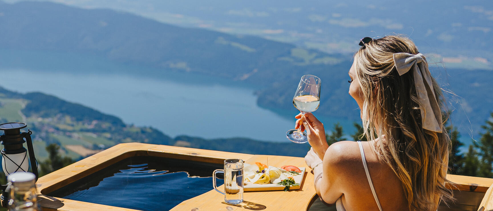 Woman with wine glass in wooden tub on mountain peak, Attersee lake and Alpine panorama in background.