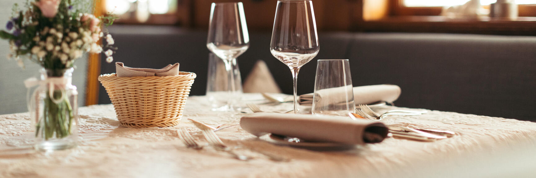 Gedeckter Tisch in einem Gasthaus mit Weingläsern, Besteck, Brotkorb und Blumenvase, Holzvertäfelung im Hintergrund.
