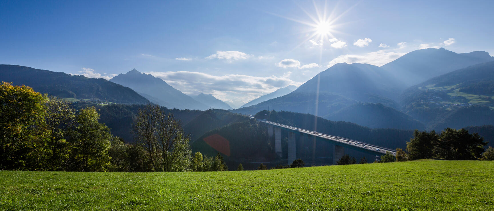 Green meadow with highway bridge and alpine panorama in background, blue sky with sun star.