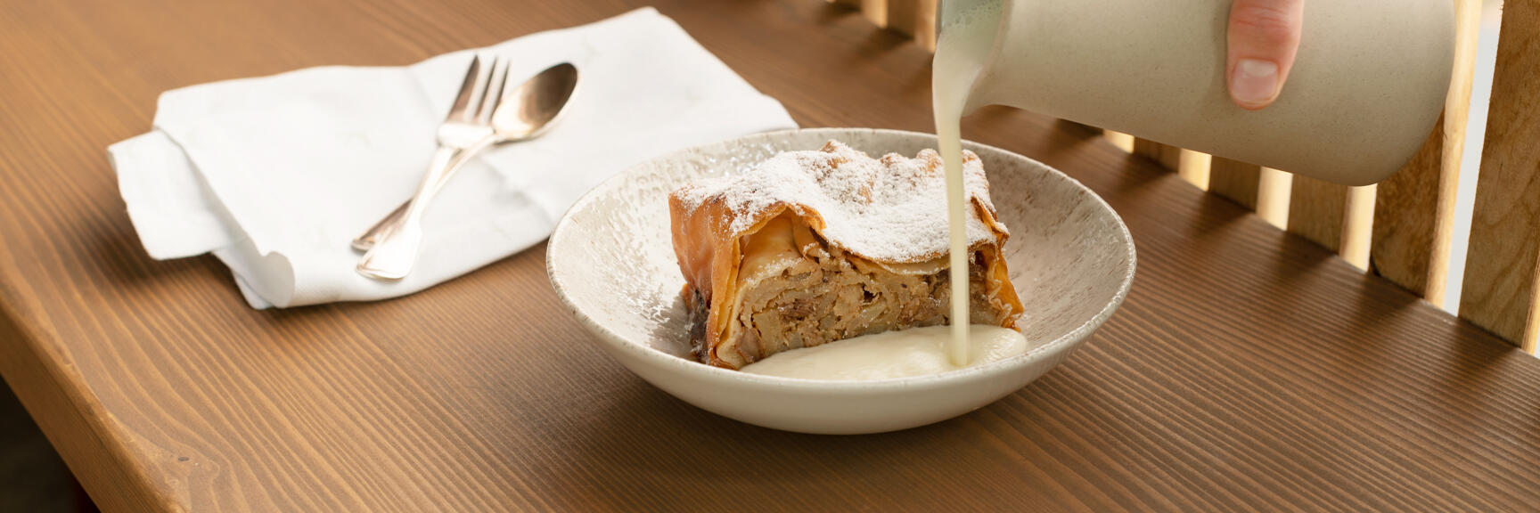 Apple strudel dusted with powdered sugar in a bowl, vanilla sauce poured from a jug.