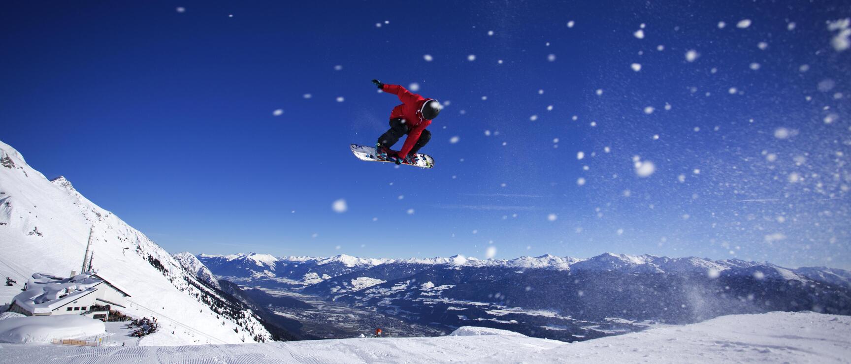 Snowboarder in red jacket jumping over snowy slope with mountain panorama and blue sky in background.