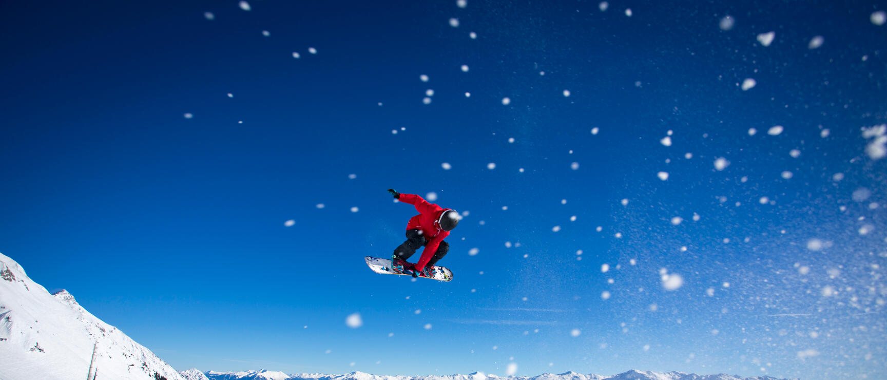 Snowboarder in red jacket jumping over snowy slope with mountain panorama and blue sky in background.