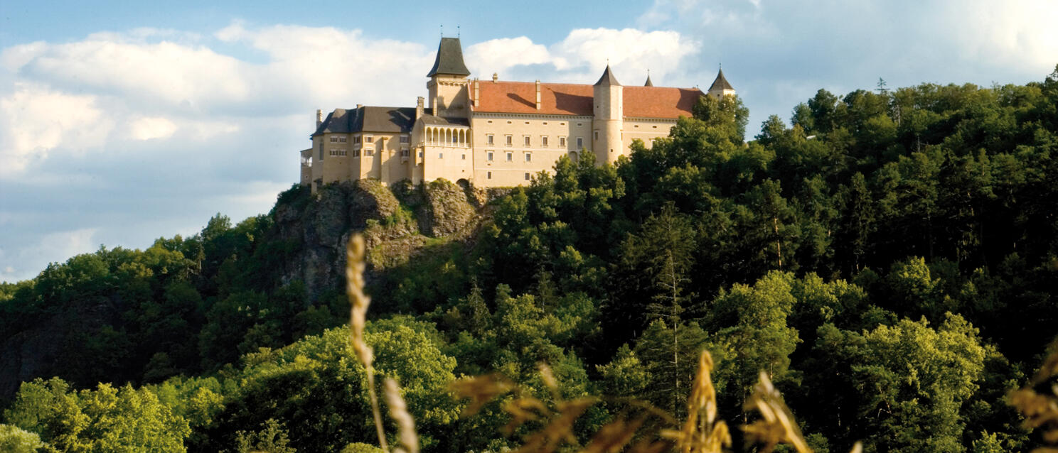 Rosenburg Castle on a forested rocky outcrop, light-coloured building with red roofs and towers, blue sky.