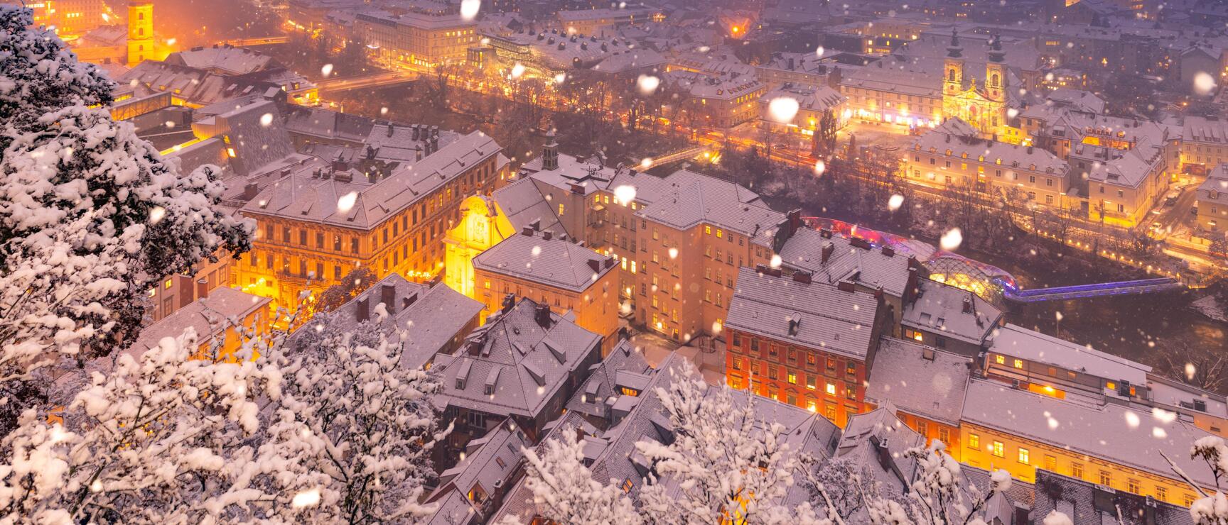 Snowfall over nightly illuminated old town of Graz, snow-covered roofs and trees in foreground.