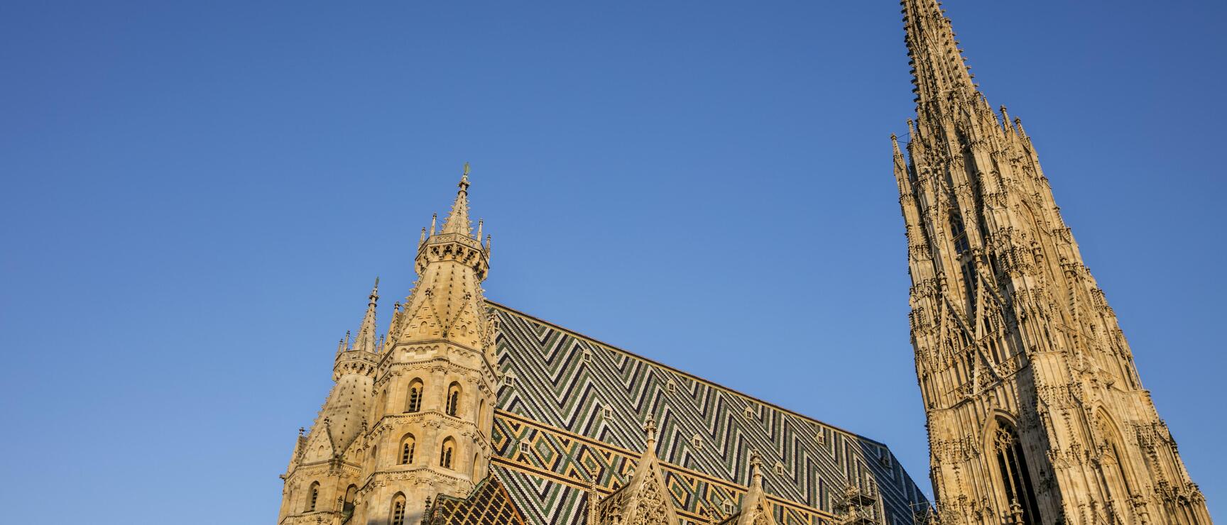 St Stephen's Cathedral in Vienna from below, Gothic towers and colourful chevron tile roof against blue sky.