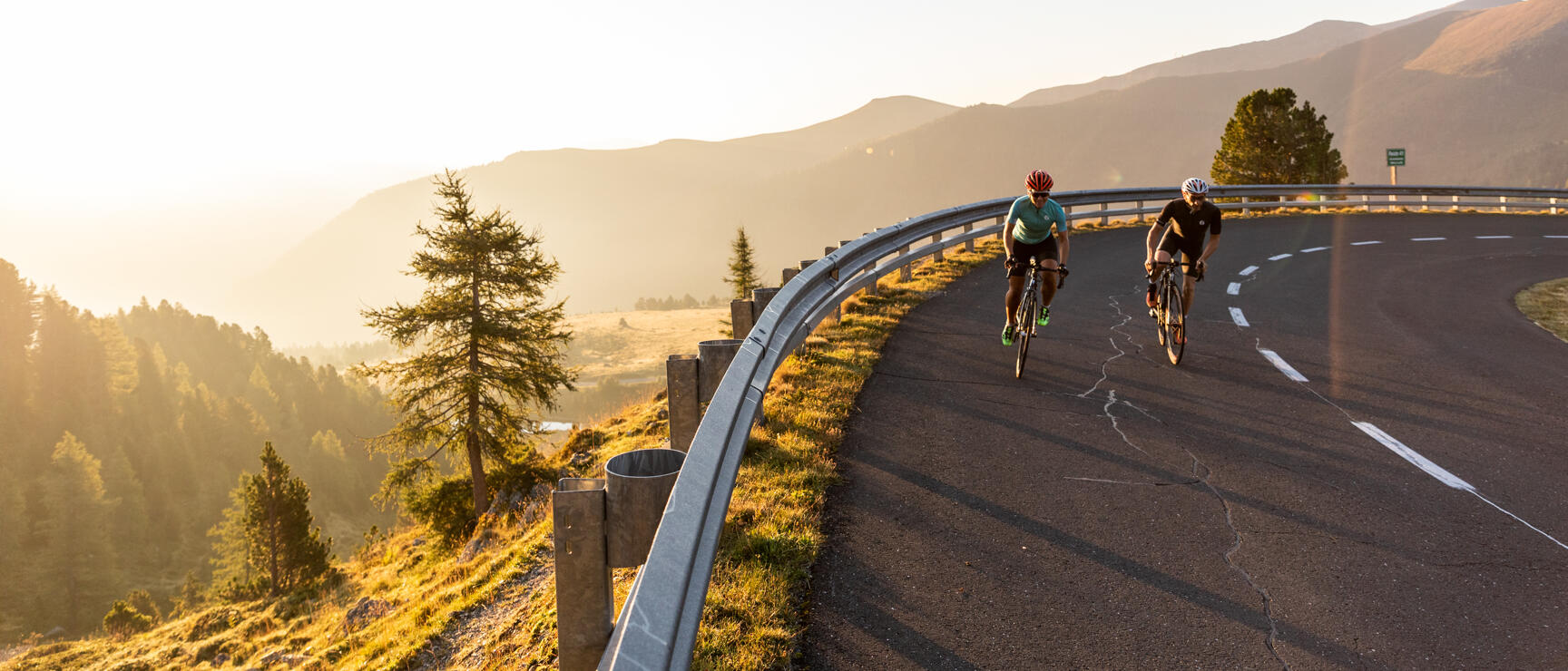 Two road cyclists on a mountain road with curve, mountain panorama and sunlight in the background.