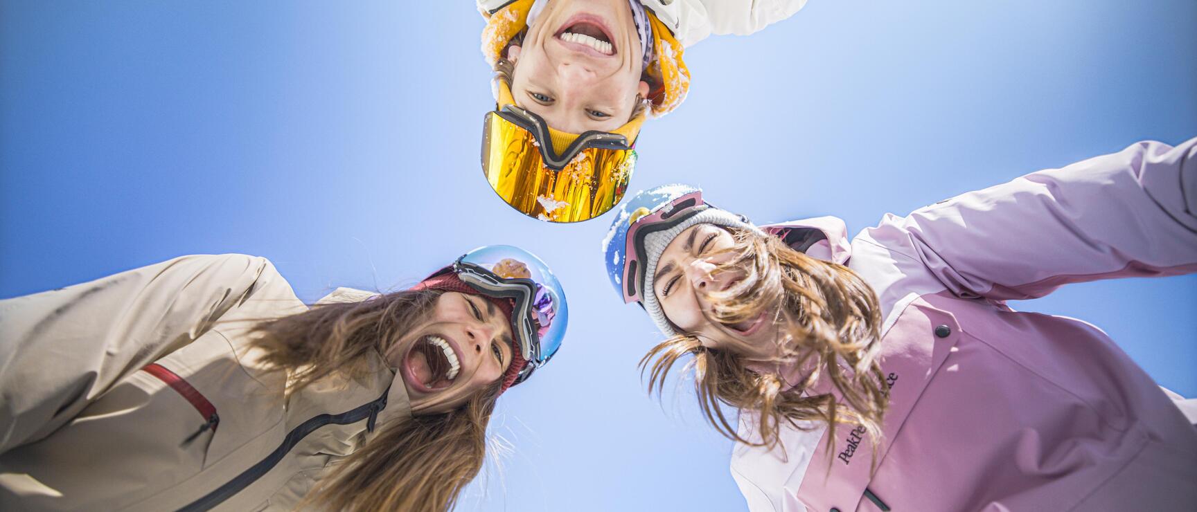 Three laughing people wearing ski helmets and goggles, photographed from below, blue sky in the background.