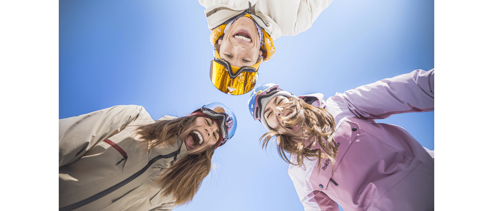 Three laughing people wearing ski helmets and goggles, photographed from below, blue sky in the background.