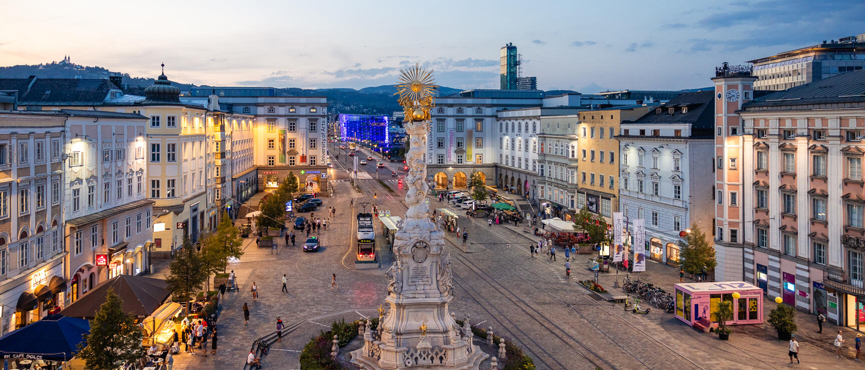 Hauptplatz Linz bei Abenddämmerung mit Dreifaltigkeitssäule, belebten Gehwegen und beleuchteten Fassaden.