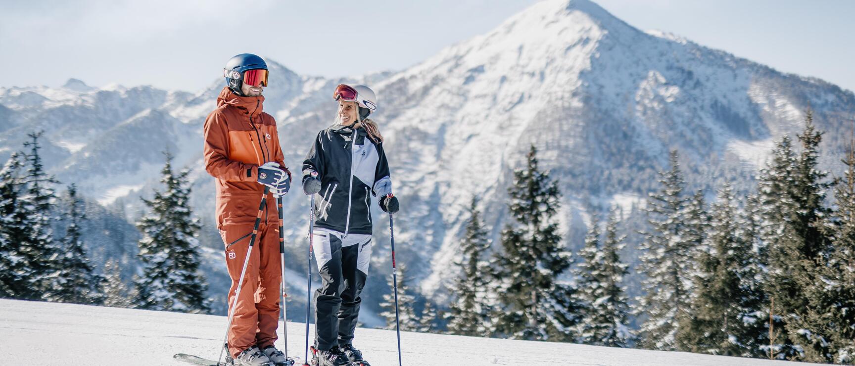 Zwei Skifahrer mit Helm und Skibrille auf verschneiter Piste, schneebedeckte Berggipfel und Tannen im Hintergrund.