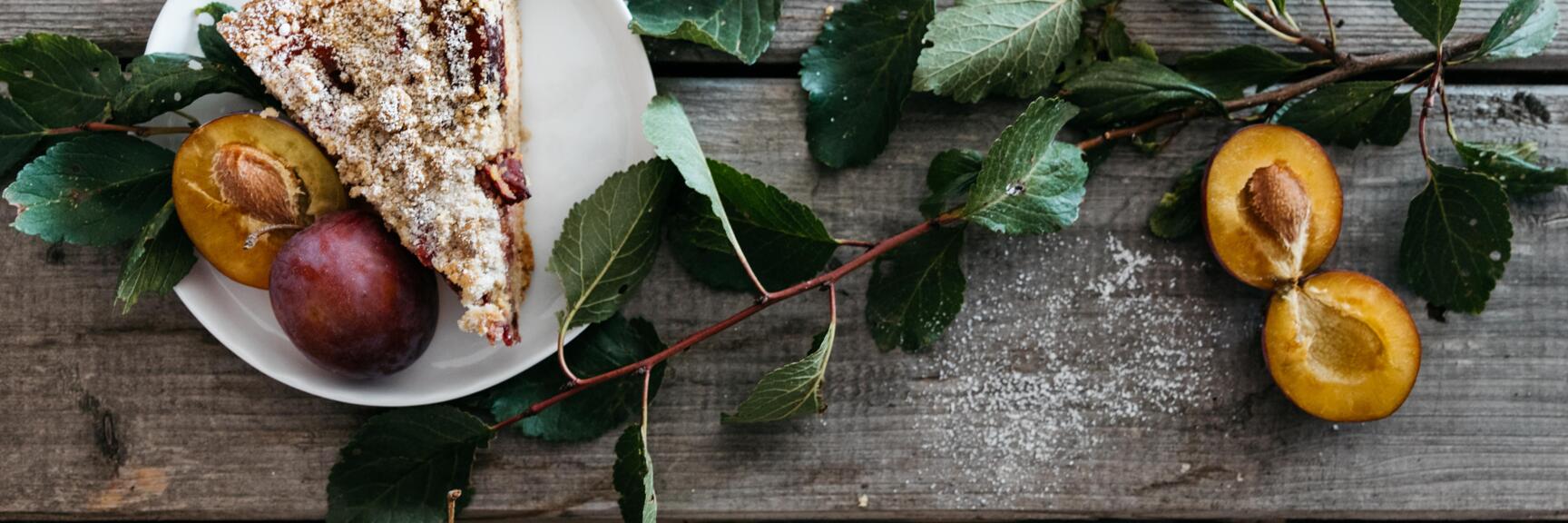 Slice of plum cake with icing sugar on plate, fresh plums and twig on rustic wooden table.