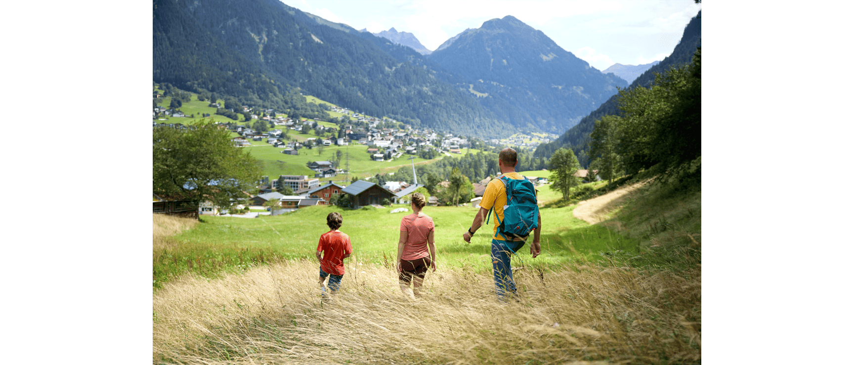 Three people hiking down a meadow with alpine village and mountain range in the background.