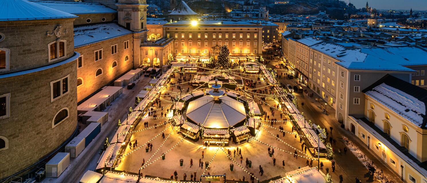 Christkindl market at Residenzplatz Salzburg, illuminated stalls in snow, cathedral and old town in background.