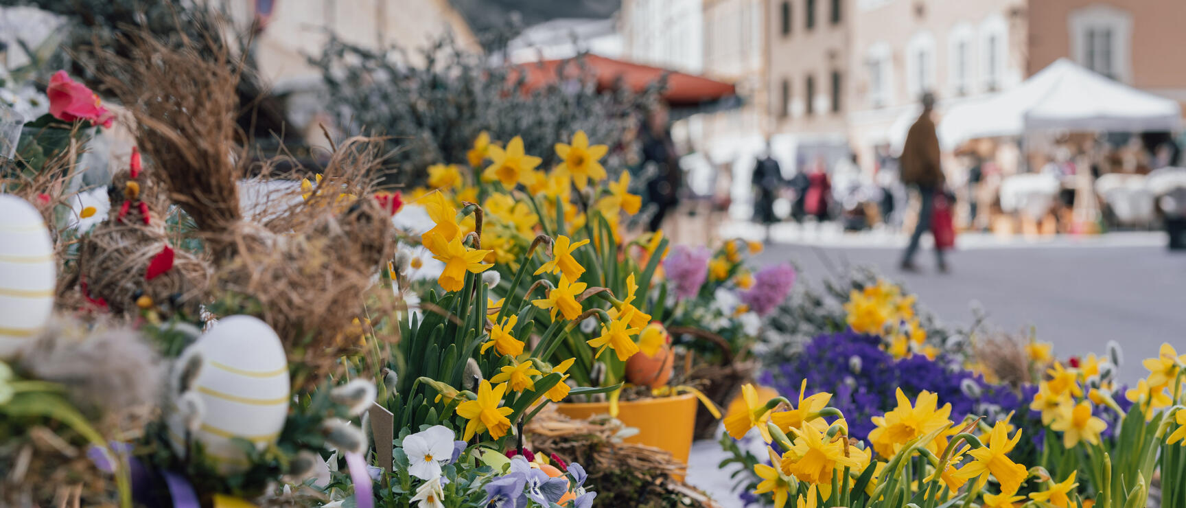 Daffodils, pansies and Easter eggs at market stall with busy town square in background.