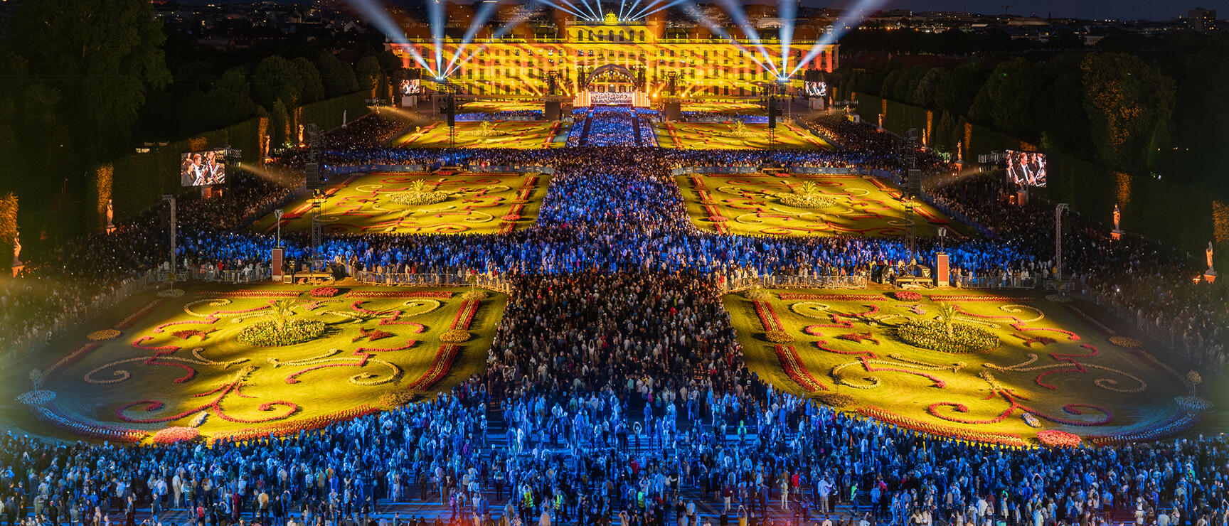 Schönbrunn Palace at night, illuminated with spotlights, crowds of people in the festively lit gardens.