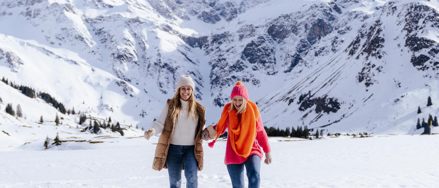 Two women in colourful winter clothing running hand in hand through snow, snow-covered Alps in background.