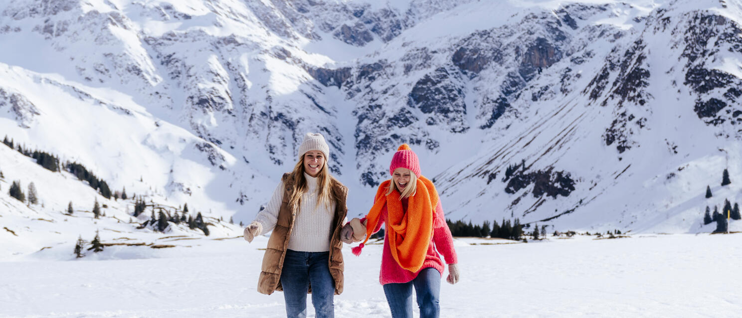 Two women in colourful winter clothing running hand in hand through snow, snow-covered Alps in background.
