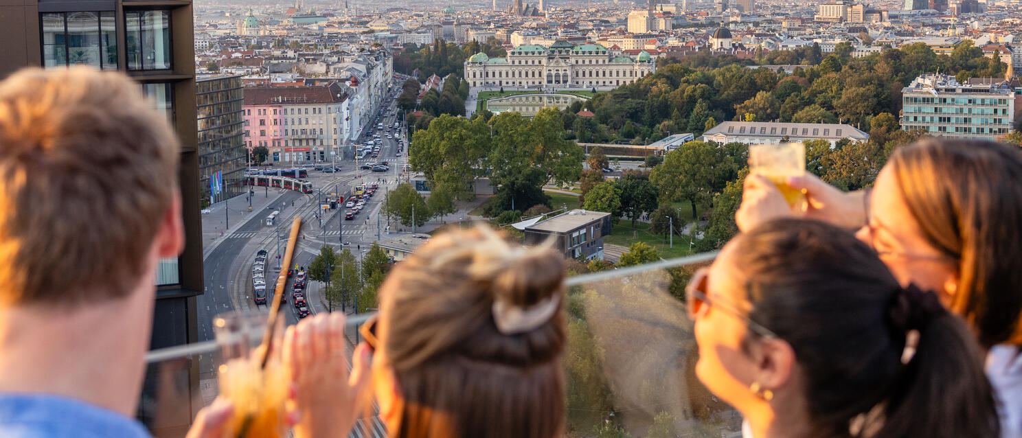 Three people with drinks on a roof terrace, view of Belvedere Palace and Vienna city skyline.