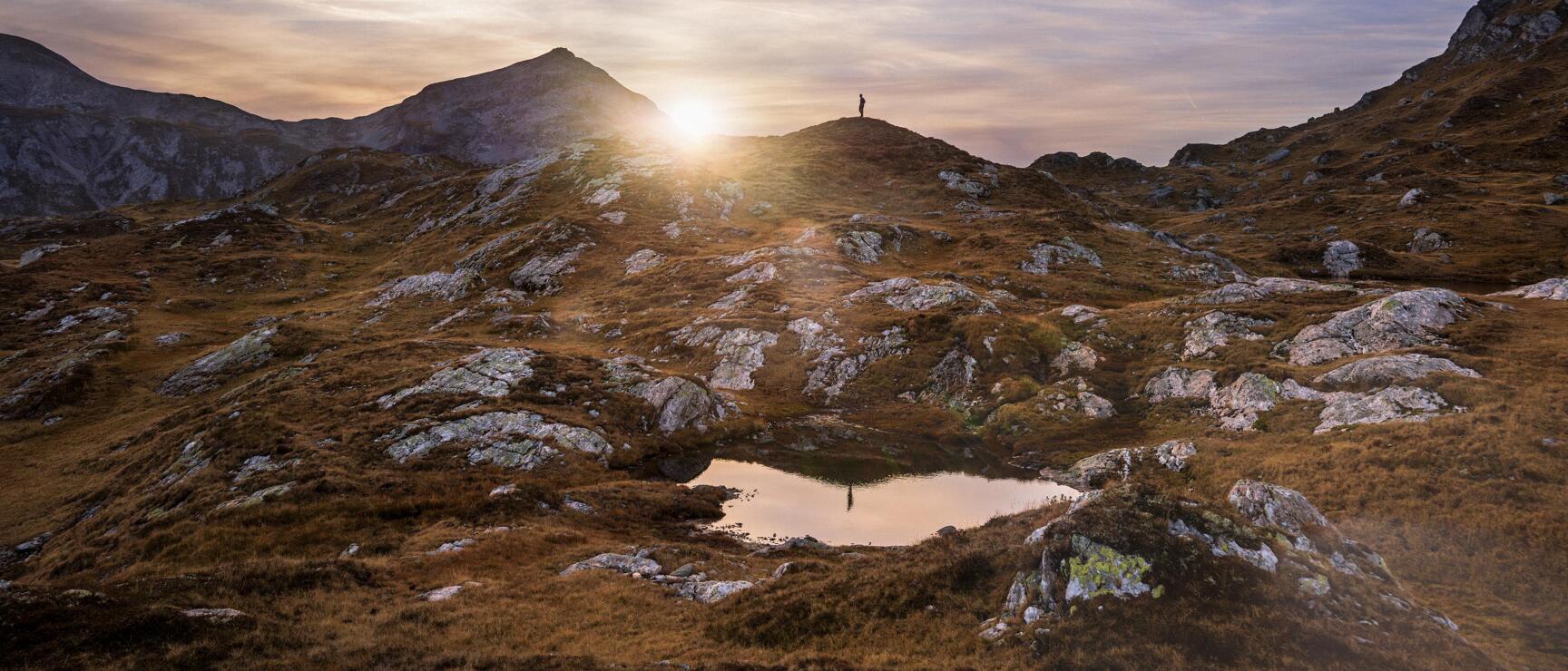 Silhouette einer Person auf Almkamm bei Sonnenuntergang, kleiner Bergsee im Vordergrund, felsige Hochalpenlandschaft.