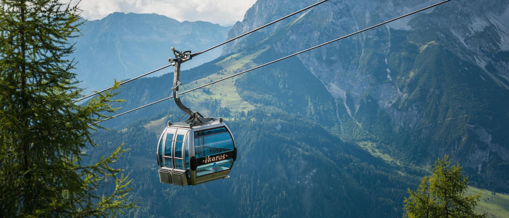 Cable car labeled 'Ikarus' on aerial tramway, surrounded by larch trees, rocky mountain backdrop.