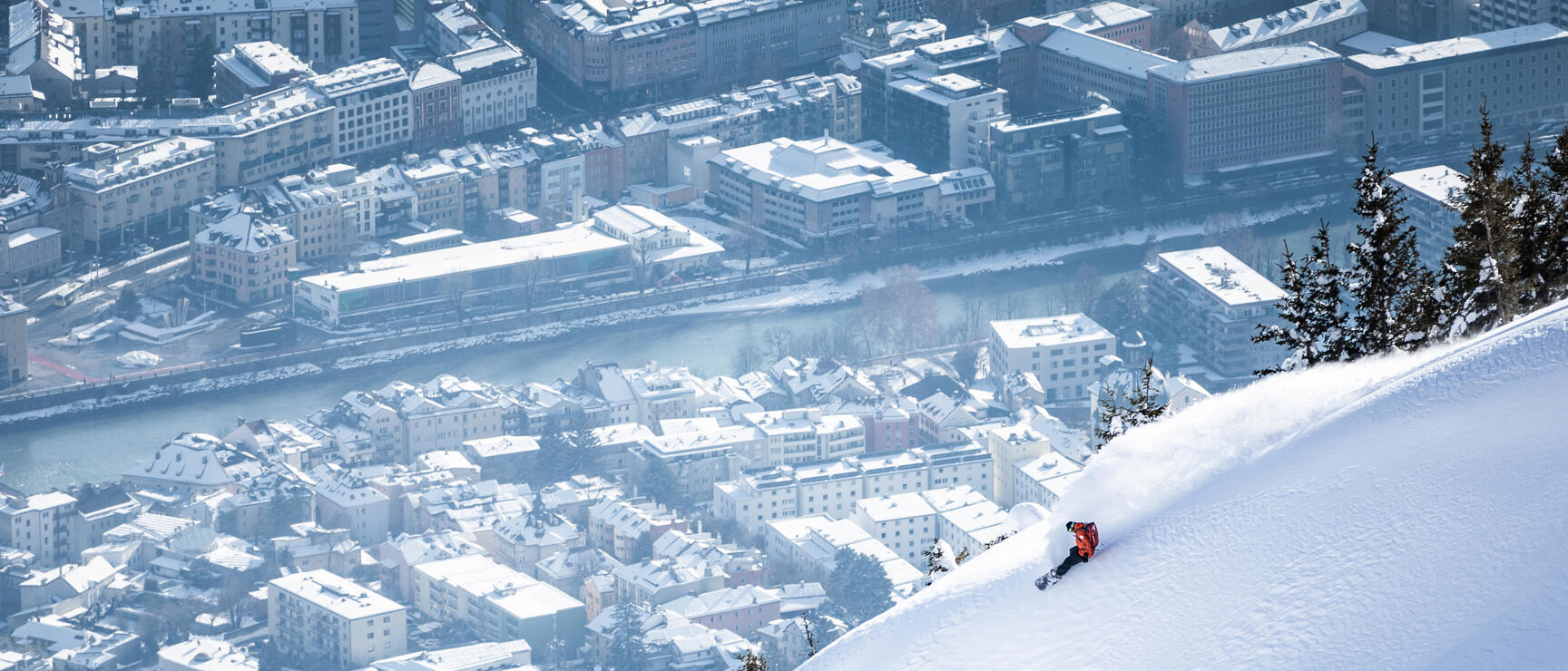 Skier in red jacket on snowy steep slope with snow-covered Innsbruck cityscape below.