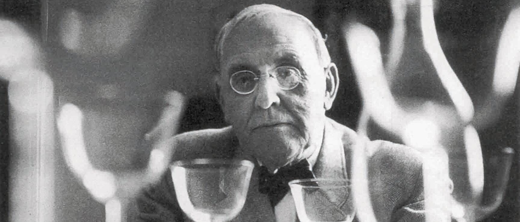 Older man with glasses and bow tie behind several wine glasses, black and white photograph.