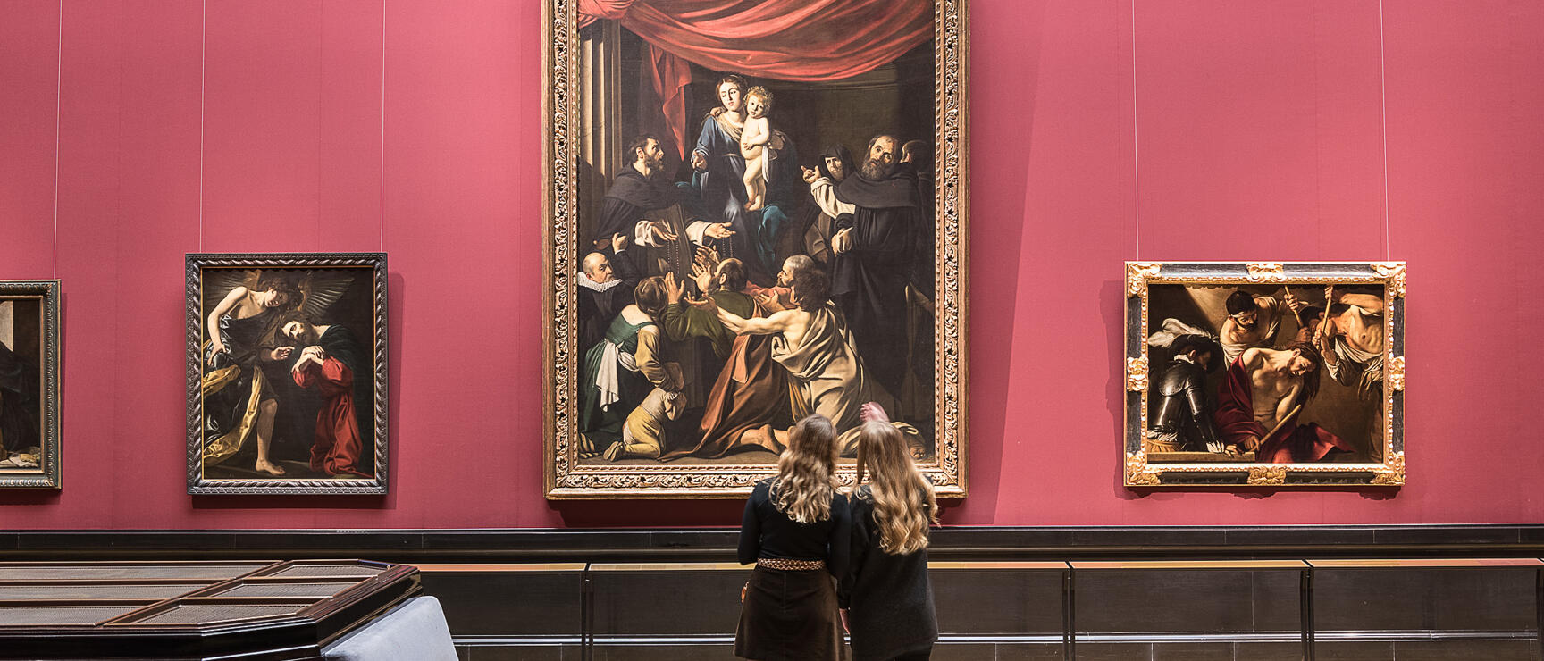 Two visitors viewing large-scale paintings on red wall at Kunsthistorisches Museum Vienna.