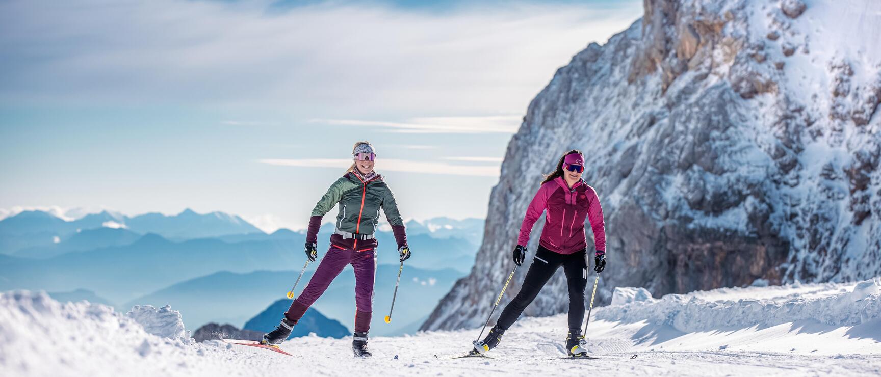 Zwei Frauen beim Skilanglauf auf schneebedecktem Plateau, Felswand und Bergpanorama im Hintergrund.