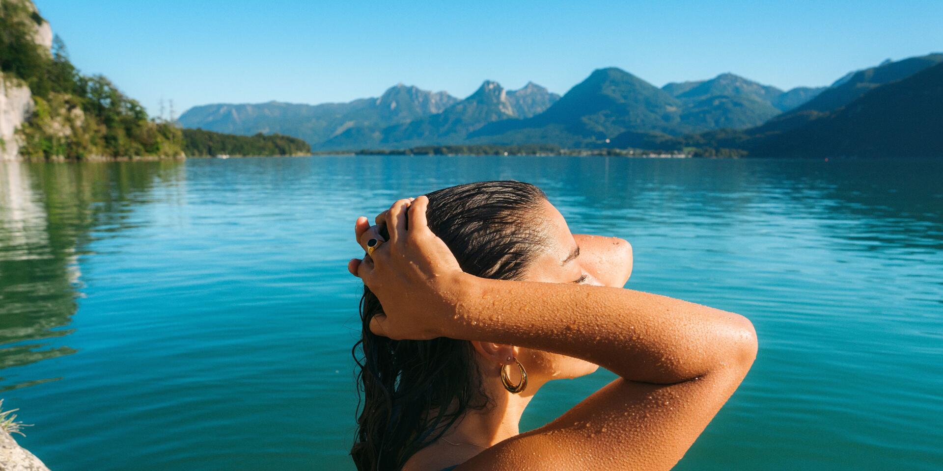 Frau im Bikini mit nassen Haaren am Seeufer, türkisblaues Wasser und Bergpanorama im Hintergrund.