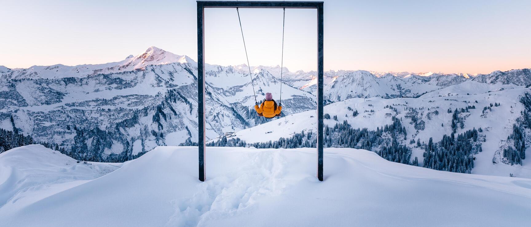 Person in yellow jacket on a mountain swing with snowy Alps and blue sky in the background.
