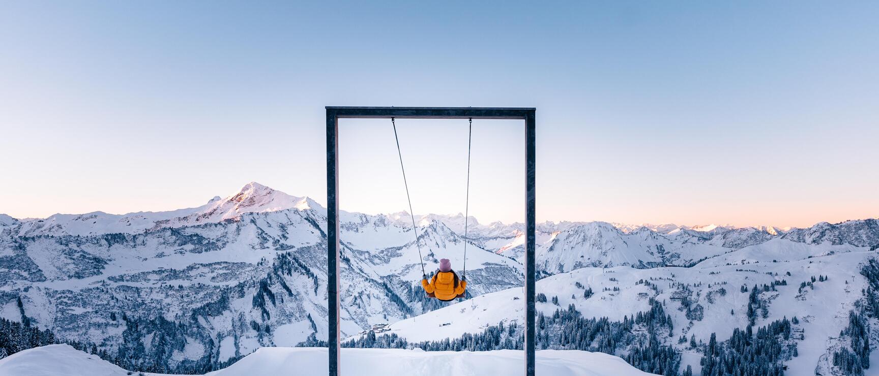 Person in gelber Jacke auf einer Bergschaukel, verschneite Alpen und blauer Himmel im Hintergrund.