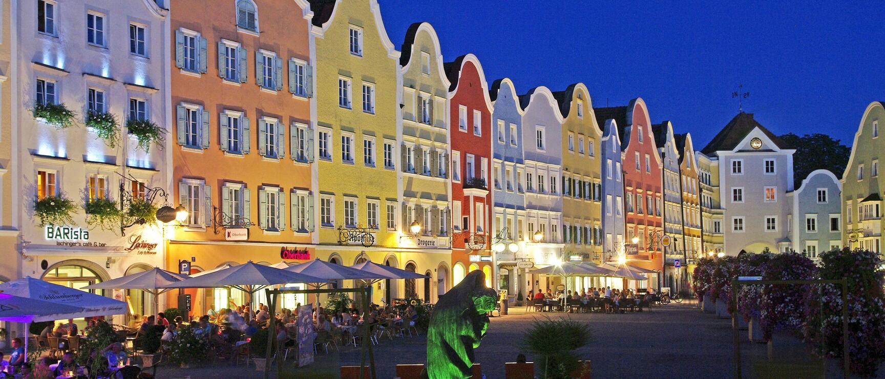 Illuminated row of houses with Baroque gables at Schärding town square at night, full moon in sky, outdoor seating area.