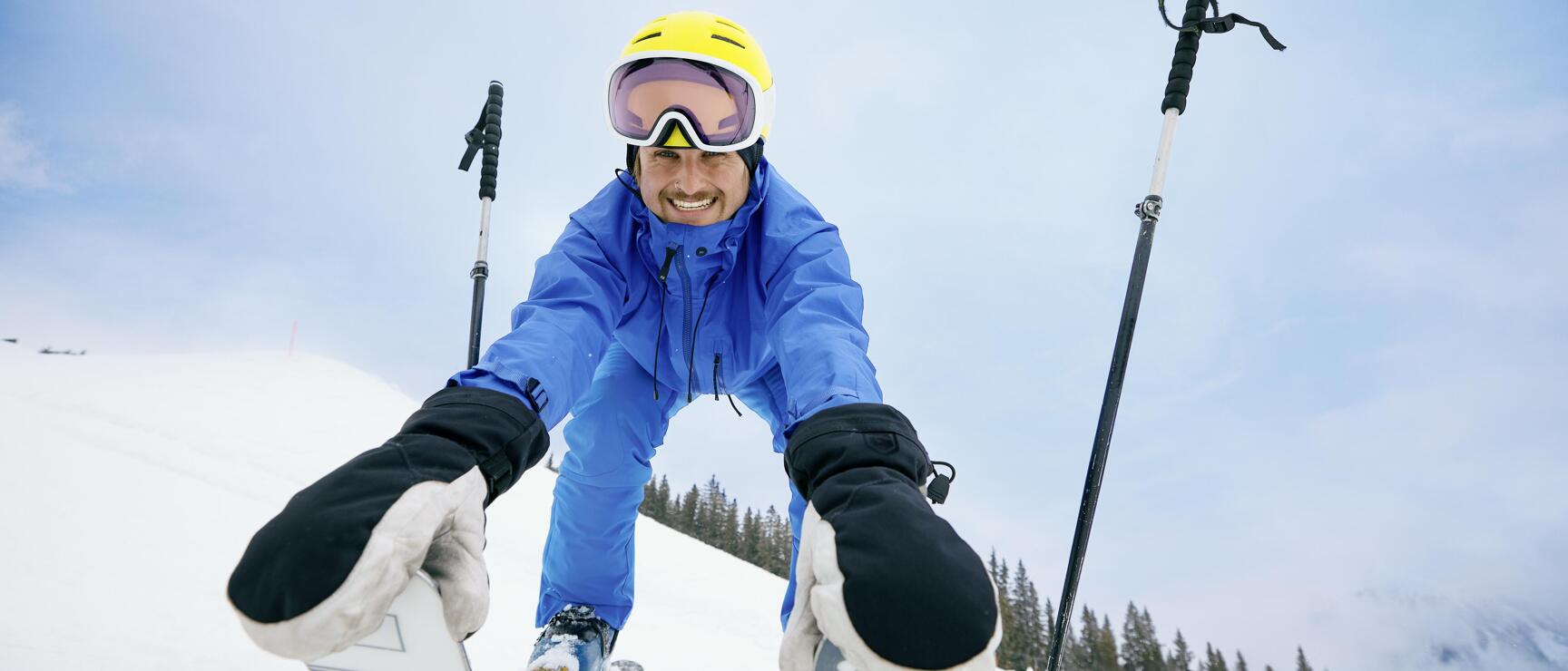 Laughing skier in blue jacket and yellow helmet sits in the snow, ski poles beside him, forest in the background.