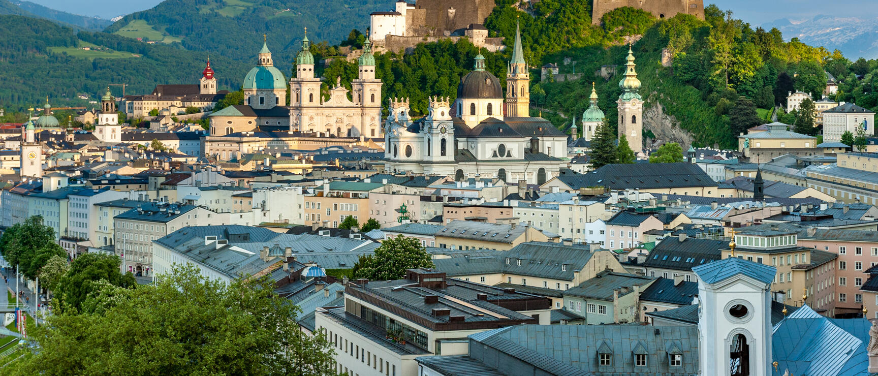 Panoramic view of Salzburg's old town with cathedral, church spires and Hohensalzburg Fortress on a wooded hill.