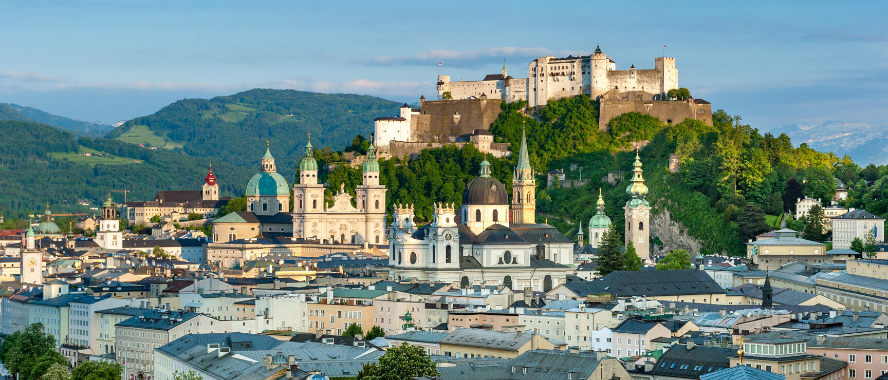 Panoramic view of Salzburg's old town with cathedral, church spires and Hohensalzburg Fortress on a wooded hill.