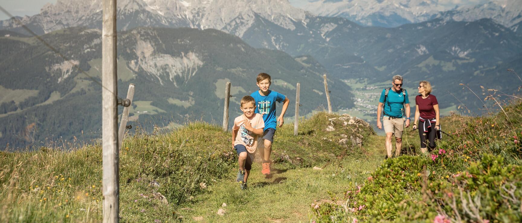 Two children running on a mountain trail with two adults following, snow-capped Alpine peaks in the background.