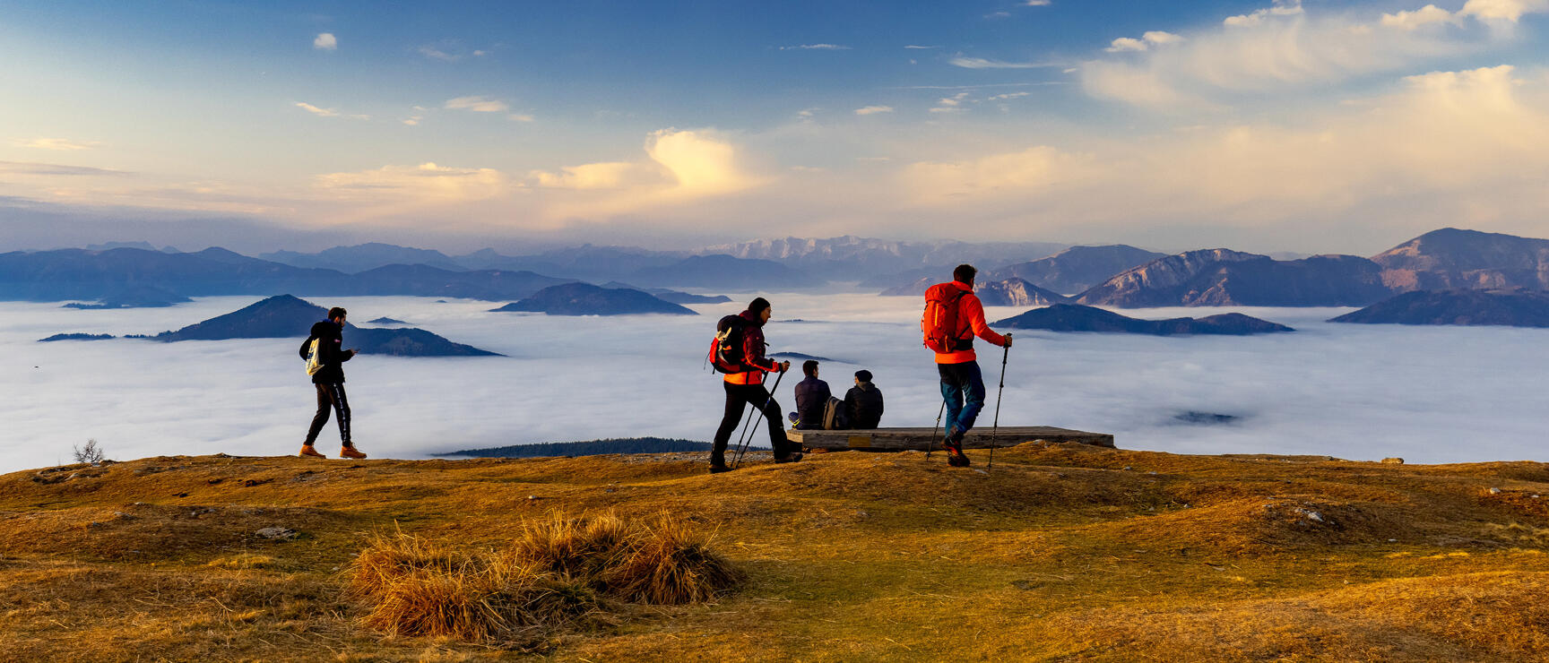 Hiking group on a mountain peak above a sea of clouds, mountain range in the background, blue sky.