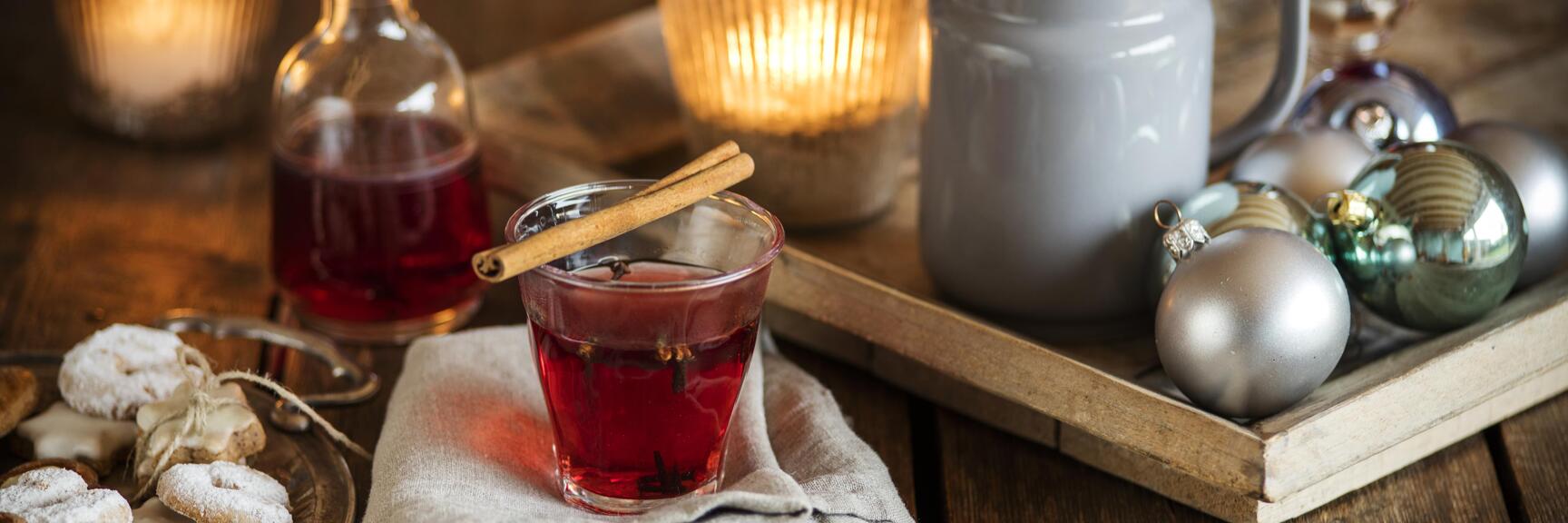 Glühwein mit Zimtstange im Glas, Weihnachtskekse und Christbaumkugeln auf einem Holztisch mit Kerzen.