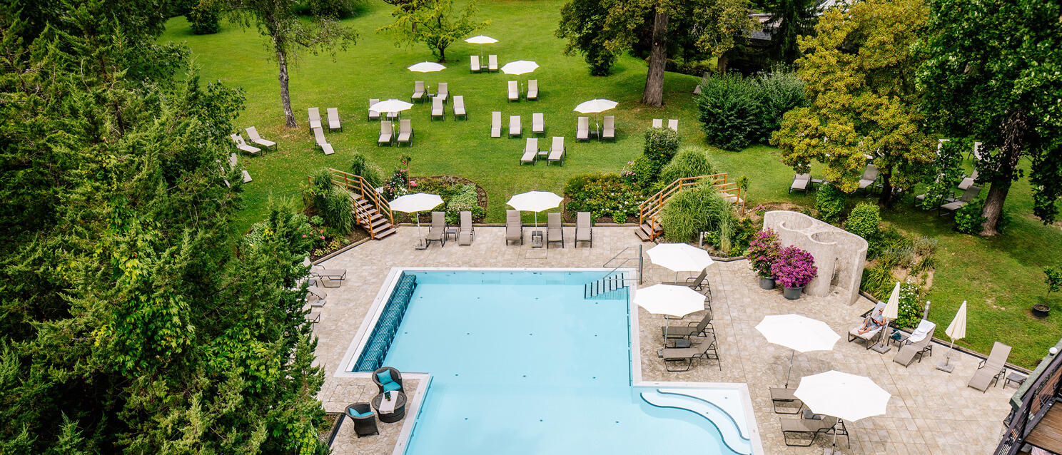 Aerial view of outdoor swimming pool with loungers and sun umbrellas, surrounded by trees and green areas.