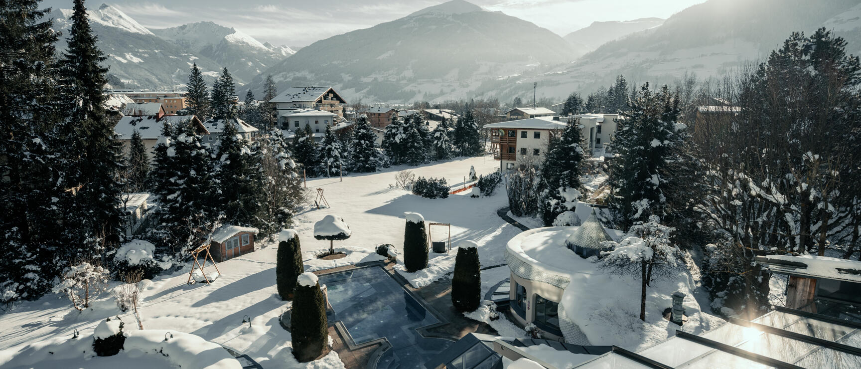 Snow-covered hotel garden with outdoor pool and circular building, alpine village and mountain panorama in the background.