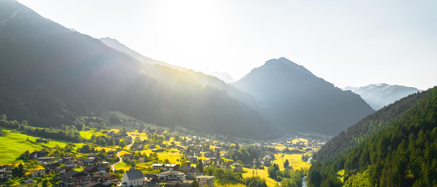 Aerial view of an alpine village with church, green meadows, river and mountains in backlighting.