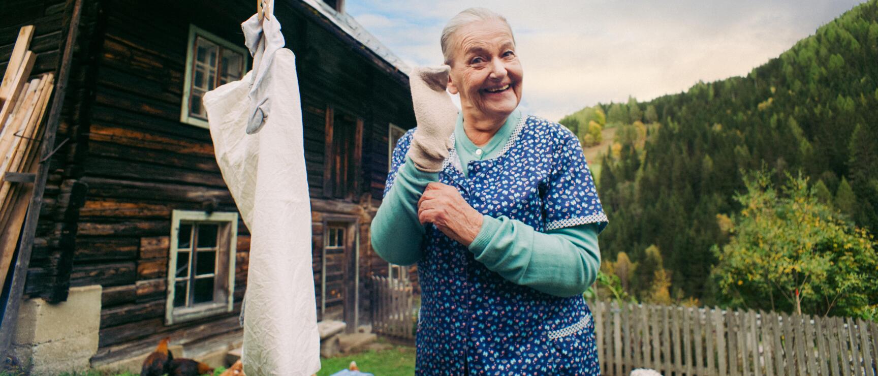 Elderly woman hanging laundry on a clothesline in front of a wooden farmhouse, chickens and forest in the background.