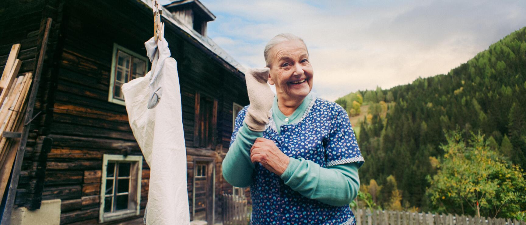 Ältere Frau hängt Wäsche auf einer Leine vor einem Holzbauernhaus auf, Hühner und Wald im Hintergrund.
