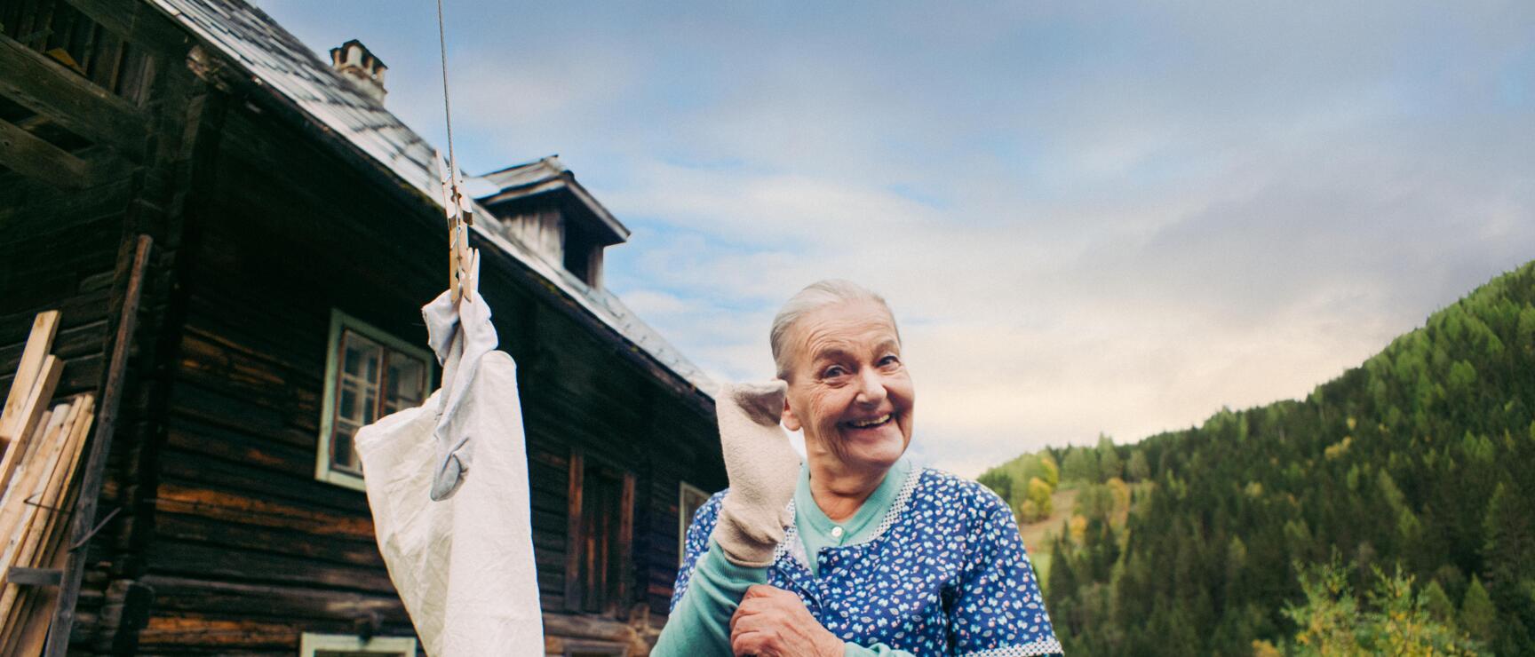 Elderly woman hanging laundry on a clothesline in front of a wooden farmhouse, chickens and forest in the background.