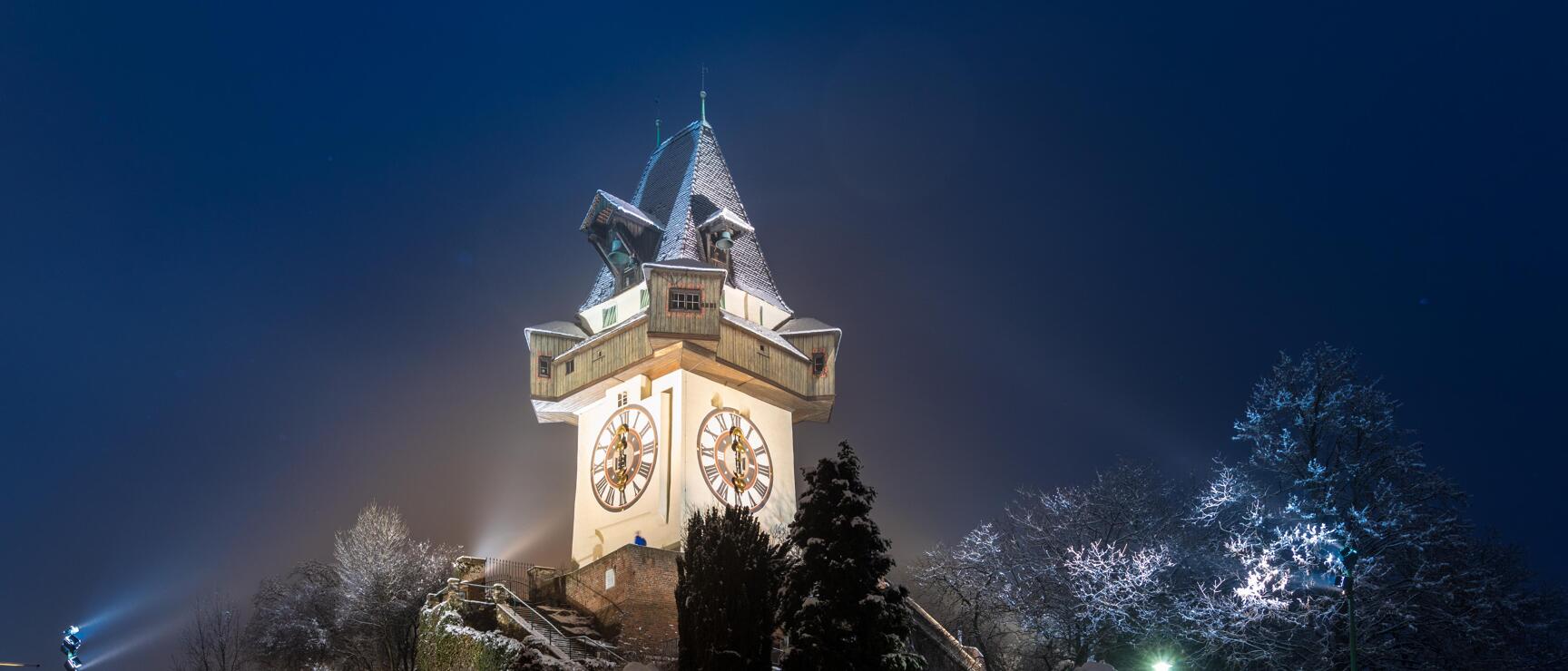 Beleuchteter Grazer Uhrturm bei Nacht im Winter, verschneite Bäume und Schlossberg rundherum.
