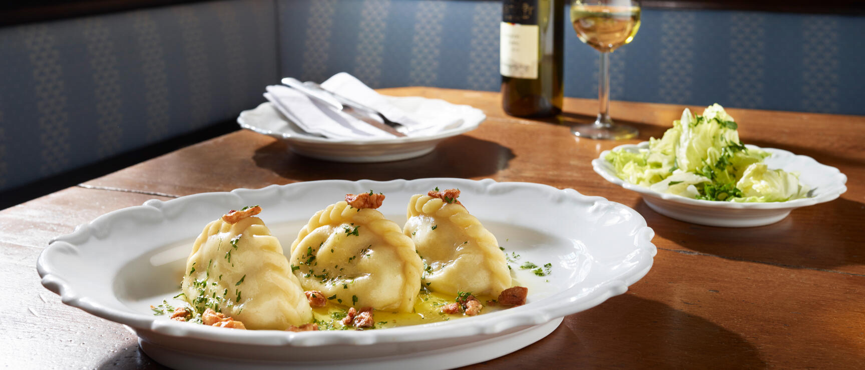 Cheese maultaschen with butter and bacon on white plate, salad and white wine in background, rustic wooden interior.
