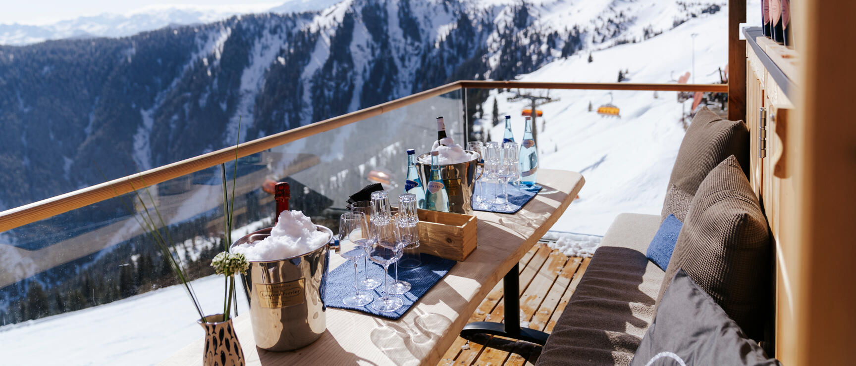 Mountain hut sun terrace with champagne glasses and ice bucket, snow-covered mountain panorama in the background.