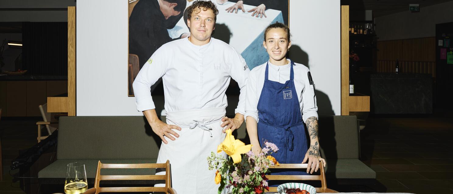 Two chefs in white chef jackets standing behind a laid table with food and flowers in front of a painting.
