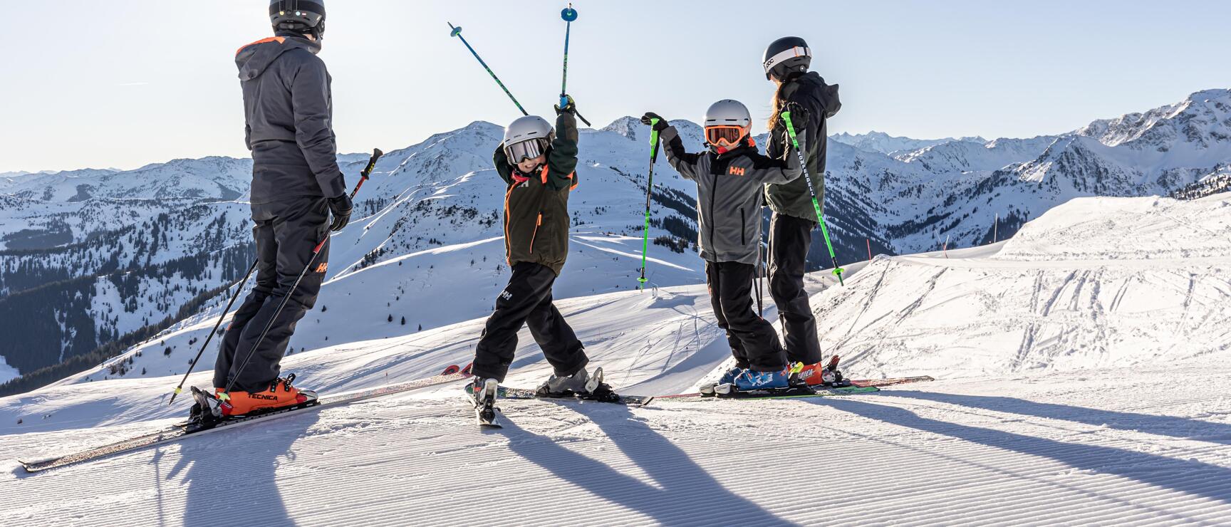 Four skiers wearing helmets on a slope with snow-covered mountains in the background and blue sky